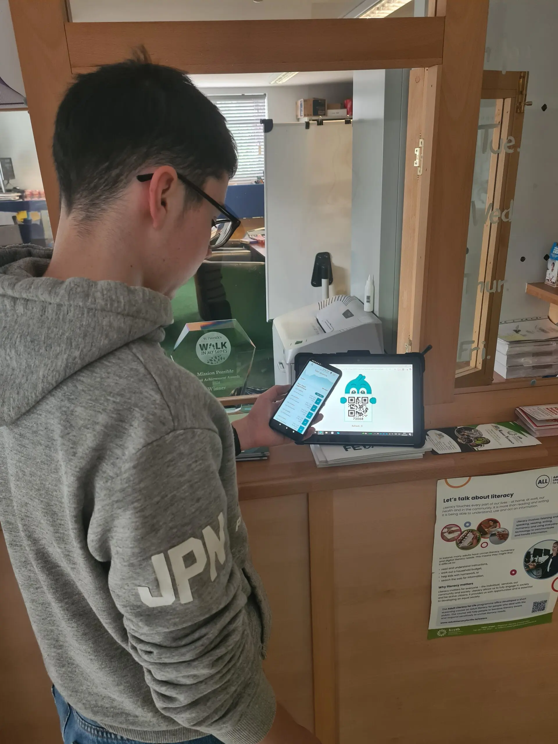 20250624_103548 Male teenager standing with a phone in front of an iPad showing a QR code